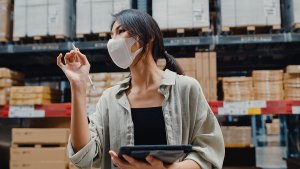 Businesswoman manager wearing face mask looking for goods using digital tablet checking inventory levels stand in retail shopping center. Wholesale Distribution, Logistics, Packages ready for shipment.